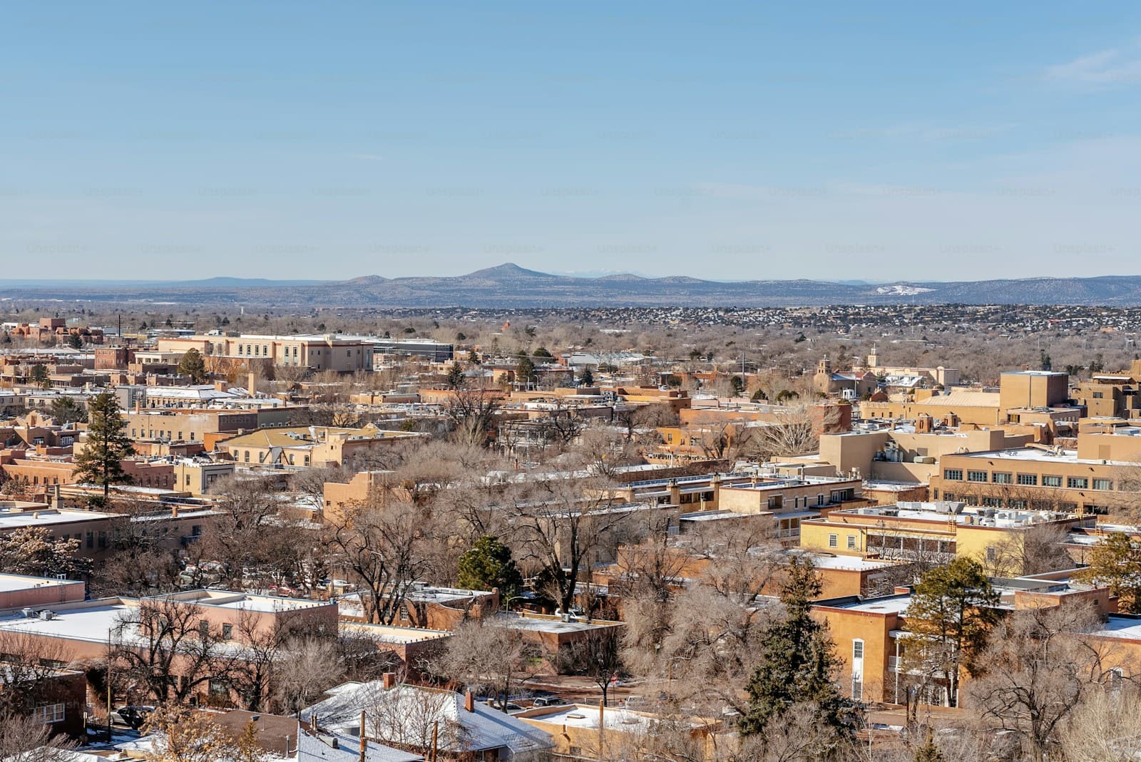 Aerial view of a southwestern city with buildings, bare trees, and mountains in the distance under blue sky